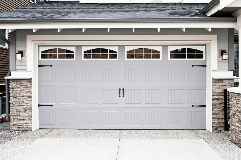Exterior view of a modern white carriage-house style garage door with decorative black hardware and arched glass windows panels installed on a residential home in Crofton
